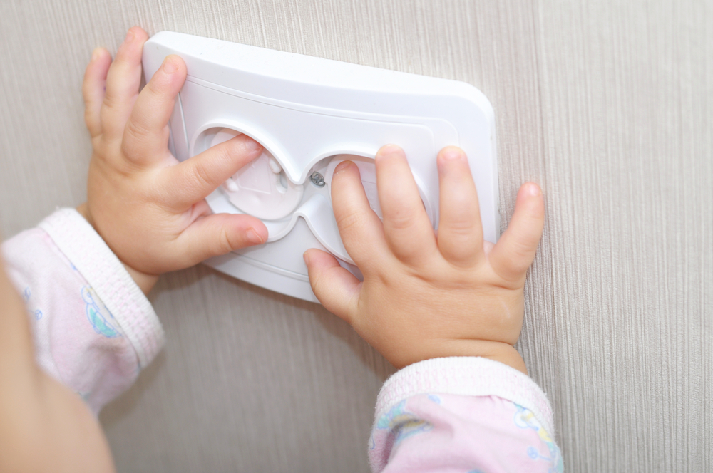 Ein Kind berührt mit seinen kleinen Fingern die Steckdose, die mit einer Plastikkappe gesichert ist.