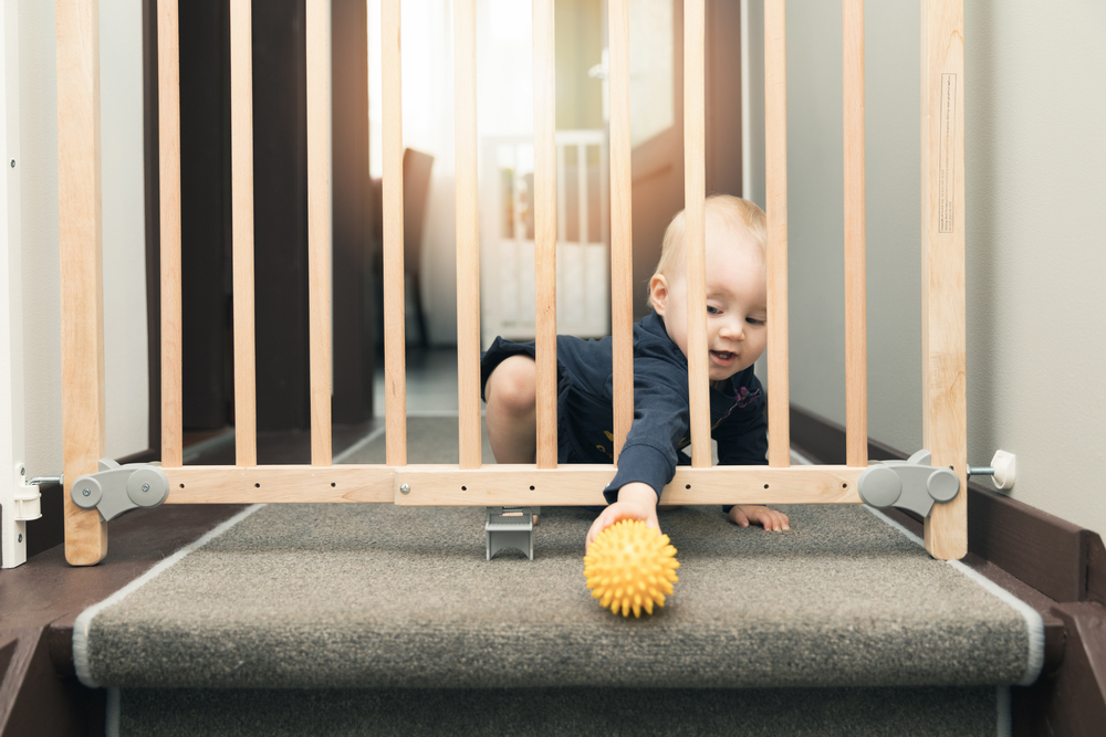 Ein Kind spielt mit einem gelben Gummiball oben auf der Treppe, befindet sich aber hinter einem Holzgeländer, um zu verhindern, dass es die Treppe hinunterfällt.